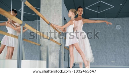 Woman teacher helps little ballerina do raise up leg leaning on wooden barre near large mirror in light studio at dance lesson