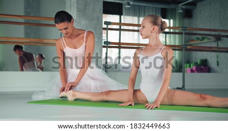 Woman teacher helps preteen girl sitting in splits stretch foot on green floor mat in brightly lit studio at ballet dance lesson