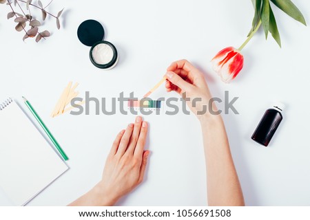 Woman testing cosmetics by using litmus paper and scale. Top view of female hands measuring of pH level in cosmetics among tulip, eucalyptus branch and notepad with pencil on white background