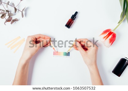 Woman testing cosmetics by using litmus papers and scale. Top view of female hands measuring of pH level among tulip, eucalyptus branch on white background, flat lay composition.