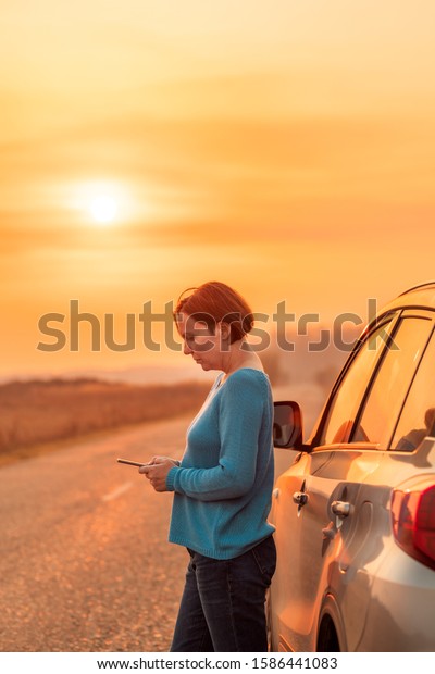 Woman\
text messaging roadside assistance help after car broke down on\
countryside road in autumn sunset, selective\
focus