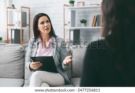 A woman therapist, dressed in a gray blazer and white pants, sits on a couch in an office setting. She is talking to her patient, who is sitting in front of her. Therapist has a clipboard in her hands
