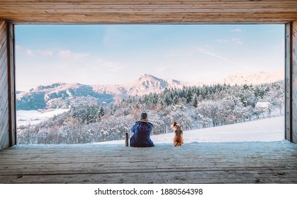 Woman with thermos flask sitting with her beagle dog in the big wooden hangar with a huge panoramic window and looking on the snowy mountain landscape. Human and pets relatives concept image.  - Powered by Shutterstock