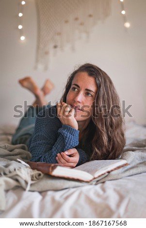 Woman thinking about what to write in her diary, whilst lying on her bed with fairy lights in the background.