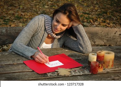 woman thoughtfully smiling, leaning on the arm, writing word LOVE on white and red paper on wooden table with candles