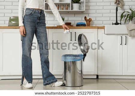 Woman throwing crumpled paper into trash bin with foot pedal in kitchen