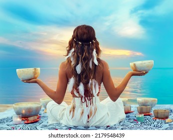 Woman with Tibetan Singing Bowls. Relaxation and Meditation at Sunset Beach. Sound Healing Therapy. Peaceful Women Silhouette practicing Yoga over Sunrise Sky
