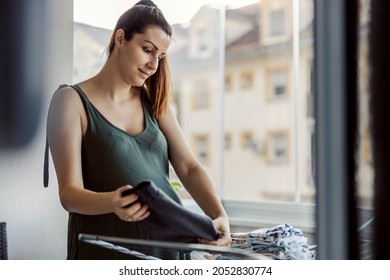 A Woman Is Tidying Up A Messy House. A Thoughtful Young Woman With A Ponytail Neatly Prepared Dry And Clean Laundry During The Day On The Terrace. House Cleaning, Housekeeping, House Chores