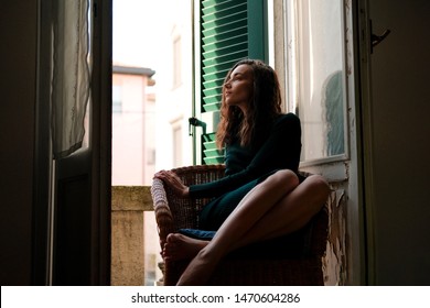 Woman In Tight Knit Dress Sitting In Wicker Chair On A Balcony With Green Wooden Shutters In Traditional Italian Hotel. Female Solo Travel Concept.