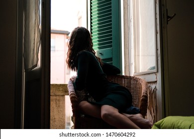 Woman In Tight Knit Dress Sitting In Wicker Chair On A Balcony With Green Wooden Shutters In Traditional Italian Hotel. Female Solo Travel Concept.