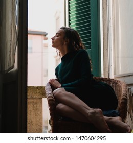 Woman In Tight Knit Dress Sitting In Wicker Chair On A Balcony With Green Wooden Shutters In Traditional Italian Hotel. Female Solo Travel Concept.