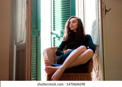 Woman In Tight Knit Dress Sitting In Wicker Chair On A Balcony With Green Wooden Shutters In Traditional Italian Hotel. Female Solo Travel Concept.