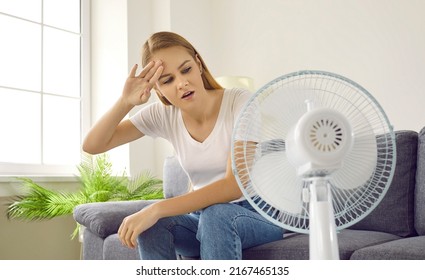 Woman Tired Of Sweltering Summertime Heat. Young Girl Sitting In Overheated Apartment, Wiping Sweaty Forehead And Trying To Get Some Fresh Air From Electric Fan Blowing In Her Face. Hot Summer Concept