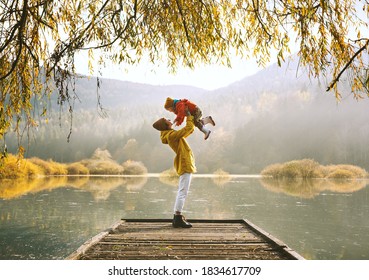 Woman with toddler on wooden pier bridge near by lake surrounded by branches of willow tree in autumn time. Mother and child together in countryside outdoors. Family in nature background.