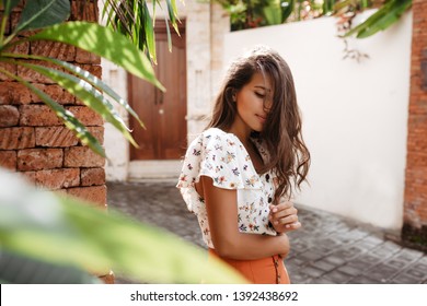 Woman touches her curly long hair. Tanned girl in bright summer outfit posing in resort town with tropical plants