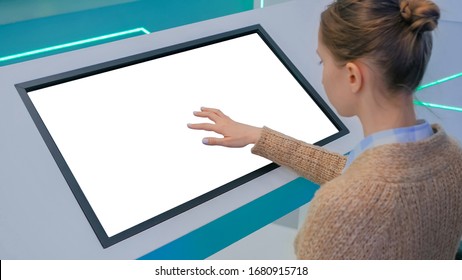Woman touching blank digital interactive white display kiosk at exhibition or museum with futuristic sci-fi interior. Mock up, copyspace, template, isolated, white screen, technology concept
