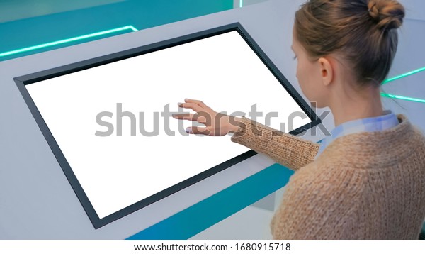 Woman touching blank digital interactive white\
display kiosk at exhibition or museum with futuristic sci-fi\
interior. Mock up, copyspace, template, isolated, white screen,\
technology concept