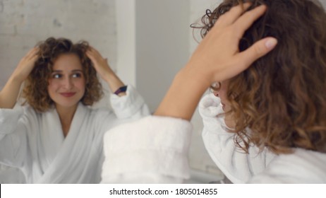 Woman touching hair and smiling looking in mirror in bathroom. Young beautiful female in bathrobe doing her hair standing in front of mirror preparing for work or date at home