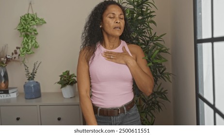 Woman touching her chest while standing in a living room, surrounded by plants and decorative items. - Powered by Shutterstock