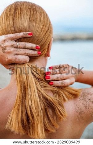Woman touching her hair with hands in sand. Rear view of red hair lady with hands with red nail polish and sand on body