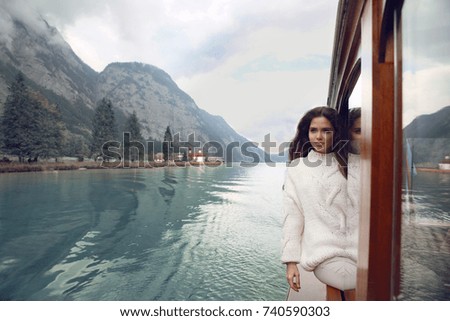 Woman tourist in cozy sweater enjoying on boat in Konigssee lake, Berchtesgaden, Germany. Young brunette traveler in warm white clothes style enjoying and relax.