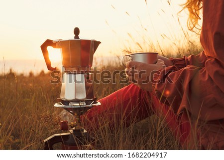 Woman tourist is drinking hot coffee from cup with coffee pot on background of sunrise meadow in mountains. Tourist makes transition, meets dawn sun. Lens flare. Camping. Lifestyle. Travel