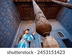 Woman tourist in ethnic dress touch wooden carved column at Inner Yard of blue mosaic palace hall in Tash Hauli of ancient city Khiva in Uzbekistan.