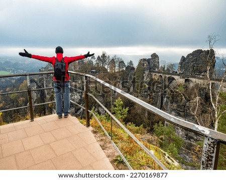 Woman tourist and hiker with rised arms is staying on view point and shouting into vally bellow. Happy woman above Bastei Bridge, Saxony park.