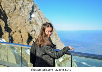 Woman tourist in the mountains.Woman stands on the observation deck high in the mountains.Swiss Alps.Rocky Mountains.Observation deck in the mountains.Woman with a backpack.Girl on vacation.happy girl - Shutterstock ID 2249458141