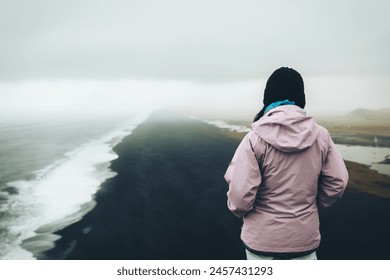 Woman tourist stand thoughtful look at atlantic ocean waves. Famous iconic cliff viewpoint over Reynisfjara black sand beach. Person Looks For Direction And Purpose On Travels - Powered by Shutterstock