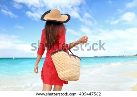 Woman tourist walking on tropical summer vacation wearing sun hat, red dress and beach bag relaxing on travel holidays. Young lady from behind in luxury fashion beachwear.