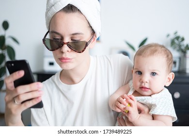 Woman With Towel Around Head In Sunglasses Holding Little Baby And Chatting, Taking Photos In Mobile Phone. Motherhood Family Lifestyle Concept. Morning Routine