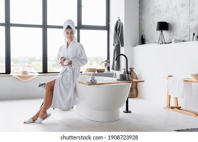 Woman in towel and bathrobe holding cosmetic cream in modern bathroom