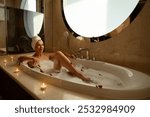 Woman with towel on her head, taking hot relaxing bath with salt and bubble bath. Beautiful young woman while enjoying bath. Lady on hotel vacation lying in bathtub. Girl while relaxing in float bath.