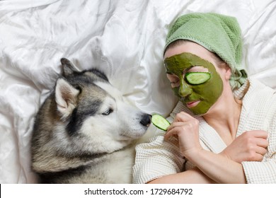 a woman in a towel on her head lies on a bed with a dog. on her face a green clay cosmetic mask with cucumbers. funny home spa with a pet. wellness concept