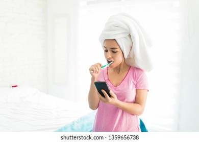 Woman with towel wrapped on head brushing teeth and using applications on mobile phone in bedroom - Powered by Shutterstock