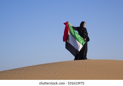 Woman in traditional Emirati dress (abaya) is holding a flag of United Arab Emirates in a desert near Dubai