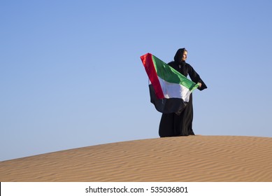 Woman in a traditional emirati dress (abaya) is holding a flag of UAE in a desert dunes