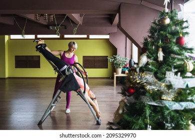 A woman with a trainer performs inversion therapy by hanging upside down on an inversion table, a therapy session is carried out in a studio near a christmas tree
