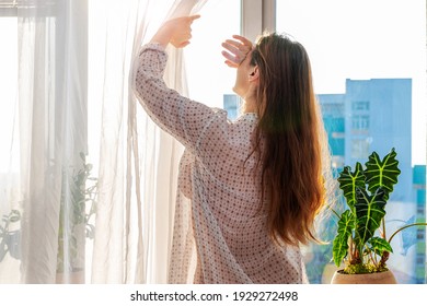 Woman In A Transparent Shirt Pulling Back The Curtain, Covered Her Face With Her Hand From The Sun's Rays, Lifestyle. Indoor Plant On The Windowsill. City Building Facade Silhouette