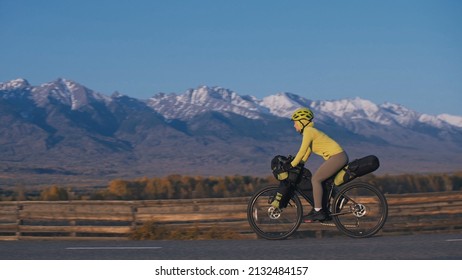 The Woman Travel On Mixed Terrain Cycle Touring With Bikepacking. The Traveler Journey With Bicycle Bags. Sport Tourism Bikepacking, Bike, Sportswear In Green Black Colors. Mountain Snow Capped.