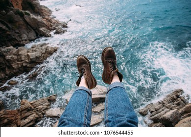 Woman Traveler In Blue Denim Jeans And Chunky Brown Leather Boots For Hiking Adventures Sits On Edge Of Cliff Or Rock Over Ocean Water, Inspiration For Travel Blog And Summer Nomad Blog Lifestyle