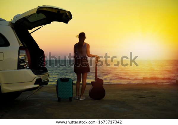 woman traveler have camera and guitar standing\
near  car and looking for view of the sea and sunset  on\
vacation.travel concept.silhouette\
style.