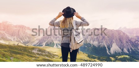 Woman traveler hipster  with backpack holding hat and looking forward  at amazing mountains and valley view. Space for text. Wearing stylish boho fall outfit. 