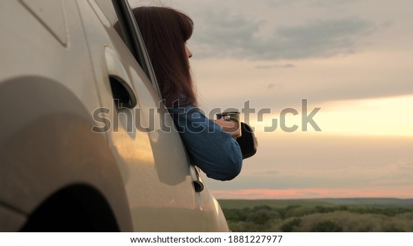 A woman traveler holds a metal mug with hot coffee\
in her hands and looks at the sunset from car window. Tourist\
driver is resting drinking tea from mug in sun. Freedom of travel\
and tourism by car.