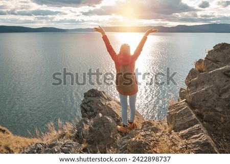 Woman traveler with open arms embracing the sky, standing on the top of the cliff with scenic lake view