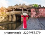 Woman traveler wearing Ao Dai Vietnamese dress sightseeing at Japanese covered bridge in Hoi An town, Vietnam. landmark and popular for tourist attractions. Vietnam and Southeast Asia travel concept