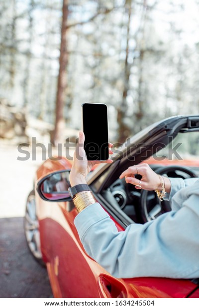 Woman traveling by car in the forest, leaning out\
of the car window and photographing with a smart phone. Phone with\
empty screen to copy\
paste