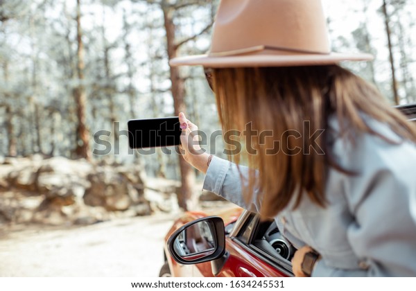 Woman traveling by car in the forest, leaning out\
of the car window and photographing with a smart phone. Phone with\
empty screen to copy\
paste