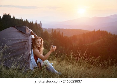 Woman traveller camping outdoors at sunset. Beautiful female sitting in tent's entrance on grass with cup of tea. Young tourist hiking, having rest in campsite. Concept of tourism in mountains. - Powered by Shutterstock